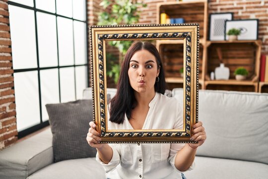 Young hispanic woman holding empty frame making fish face with mouth and squinting eyes, crazy and comical.