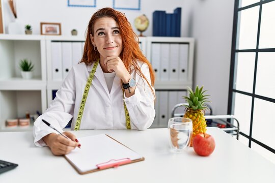 Young redhead woman nutritionist doctor at the clinic with hand on chin thinking about question, pensive expression. smiling and thoughtful face. doubt concept.