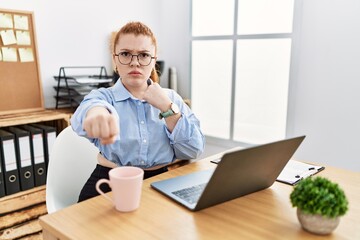 Young redhead woman working at the office using computer laptop punching fist to fight, aggressive and angry attack, threat and violence