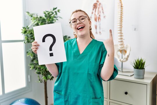 Young redhead physiotherapist woman holding question mark at the clinic smiling with an idea or question pointing finger with happy face, number one