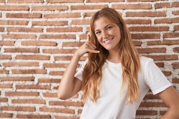 Young caucasian woman standing over bricks wall smiling doing phone gesture with hand and fingers like talking on the telephone. communicating concepts.