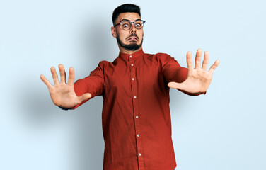 Fototapeta premium Young hispanic man with beard wearing business shirt and glasses afraid and terrified with fear expression stop gesture with hands, shouting in shock. panic concept.