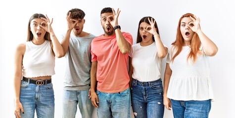 Group of young friends standing together over isolated background doing ok gesture shocked with surprised face, eye looking through fingers. unbelieving expression.