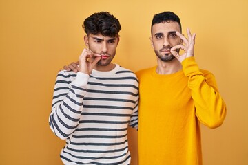 Young hispanic gay couple standing over yellow background mouth and lips shut as zip with fingers....