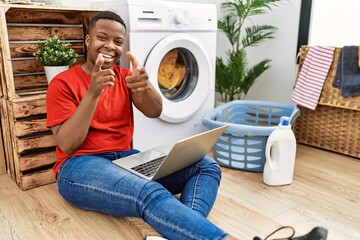 Young african man doing laundry and using computer pointing fingers to camera with happy and funny face. good energy and vibes.