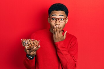 Young african american man holding bowl of chestnuts covering mouth with hand, shocked and afraid...