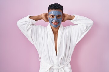 Young hispanic man wearing beauty face mask and bath robe relaxing and stretching, arms and hands behind head and neck smiling happy