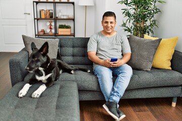 Young hispanic man using smartphone sitting on the sofa with dog at home.