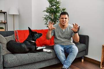Young latin man and dog sitting on the sofa at home smiling funny doing claw gesture as cat,...