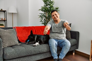 Young latin man and dog sitting on the sofa at home approving doing positive gesture with hand, thumbs up smiling and happy for success. winner gesture.
