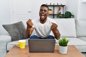 Young african man using laptop at home very happy and excited doing winner gesture with arms raised, smiling and screaming for success. celebration concept.