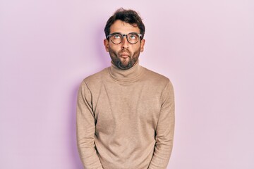 Handsome man with beard wearing turtleneck sweater and glasses making fish face with lips, crazy and comical gesture. funny expression.