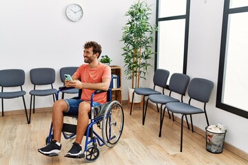 Young hispanic man using smartphone sitting on wheelchair at clinic waiting room.