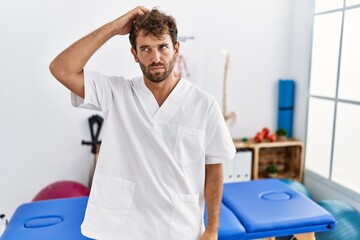 Young handsome physiotherapist man working at pain recovery clinic confuse and wondering about question. uncertain with doubt, thinking with hand on head. pensive concept.
