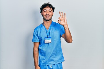 Young hispanic man wearing blue male nurse uniform showing and pointing up with fingers number three while smiling confident and happy.