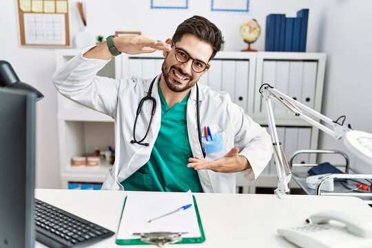 Young man with beard wearing doctor uniform and stethoscope at the clinic gesturing with hands showing big and large size sign, measure symbol. smiling looking at the camera. measuring concept.