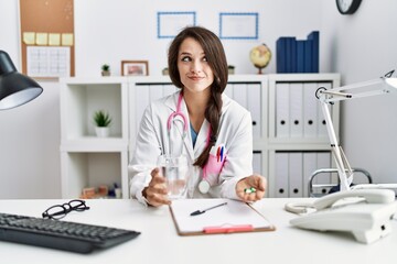 Young doctor woman holding glass of water and prescription pills smiling looking to the side and staring away thinking.