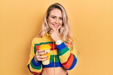 Beautiful young blonde woman drinking cup of coffee wearing headphones looking confident at the camera smiling with crossed arms and hand raised on chin. thinking positive.