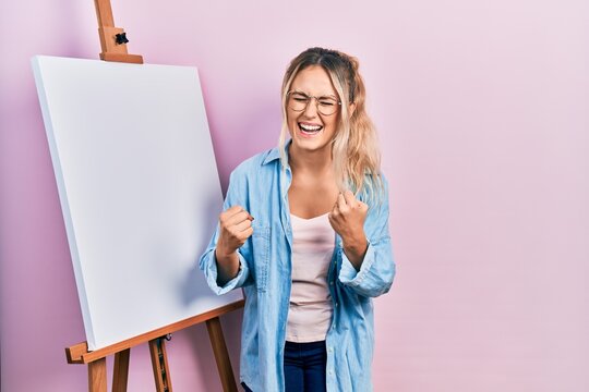 Beautiful young blonde woman standing by white painter easel stand celebrating surprised and amazed for success with arms raised and eyes closed