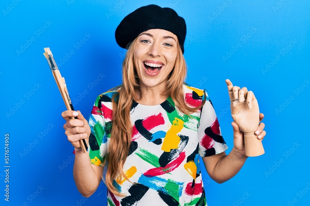 Poster Young caucasian woman artist holding small wooden manikin and paintbrushes smiling and laughing hard out loud because funny crazy joke.