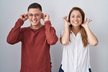Mother and son standing together over isolated background smiling pulling ears with fingers, funny...