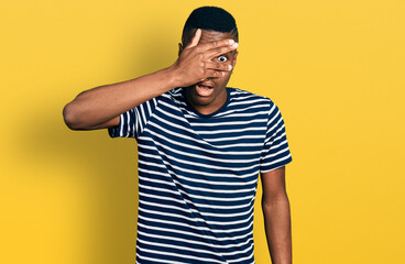 Young african american man wearing casual striped t shirt peeking in shock covering face and eyes with hand, looking through fingers afraid