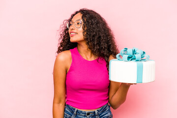 Young caucasian woman holding a cake isolated on pink background looks aside smiling, cheerful and pleasant.