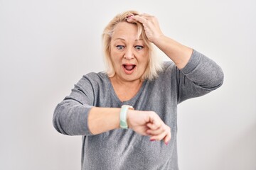 Middle age caucasian woman standing over white background looking at the watch time worried, afraid of getting late
