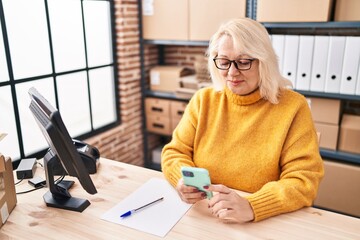 Middle age blonde woman ecommerce business worker using smartphone at office