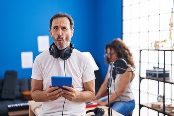 Middle age hispanic man at music studio using tablet making fish face with mouth and squinting eyes, crazy and comical.