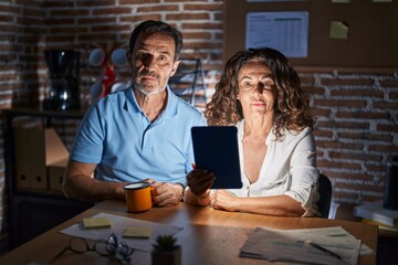 Middle age hispanic couple using touchpad sitting on the table at night relaxed with serious expression on face. simple and natural looking at the camera.