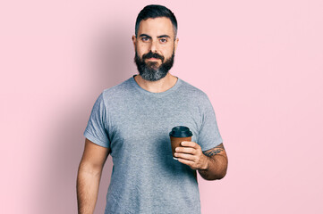 Hispanic man with beard drinking a take away cup of coffee looking positive and happy standing and smiling with a confident smile showing teeth