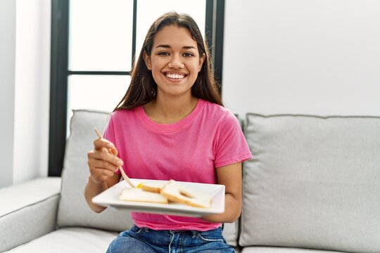 Young latin woman having lunch sitting on sofa at home