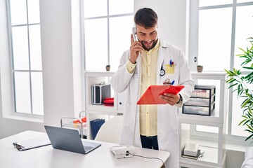 Young hispanic man wearing doctor uniform talking on the telephone at clinic