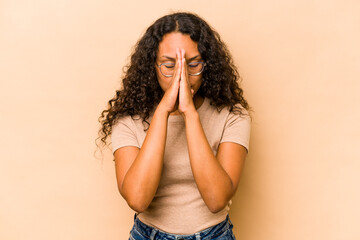 Young hispanic woman isolated on beige background holding hands in pray near mouth, feels confident.