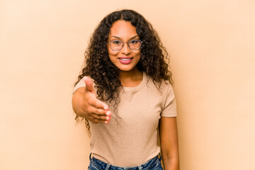 Young hispanic woman isolated on beige background stretching hand at camera in greeting gesture.