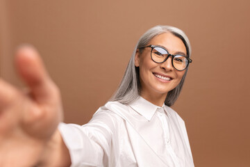 Lady wearing glasses smiling and looks at the camera isolated on beige. Cheerful senior asian woman taking selfie. POV
