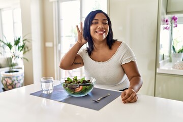 Obraz na płótnie Canvas Young hispanic woman eating healthy salad at home smiling with hand over ear listening an hearing to rumor or gossip. deafness concept.
