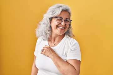 Middle age woman with grey hair standing over yellow background winking looking at the camera with...