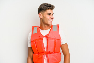 Young caucasian man wearing life jacket isolated on white background looks aside smiling, cheerful and pleasant.