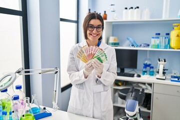 Young hispanic woman working at scientist laboratory holding money banknotes smiling with a happy...