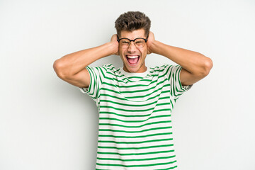 Young caucasian man isolated on white background covering ears with hands trying not to hear too loud sound.