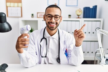 African american doctor man holding thermometer at the clinic smiling happy pointing with hand and finger to the side