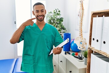 African american physiotherapist man working at pain recovery clinic smiling cheerful showing and pointing with fingers teeth and mouth. dental health concept.