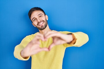 Hispanic man standing over blue background smiling in love doing heart symbol shape with hands. romantic concept.