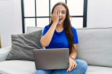 Young brunette woman using laptop at home covering one eye with hand, confident smile on face and surprise emotion.