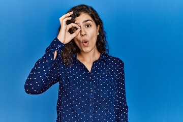 Fototapeta premium Young brunette woman with curly hair wearing casual clothes over blue background doing ok gesture shocked with surprised face, eye looking through fingers. unbelieving expression.