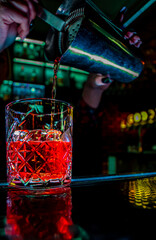 Bartender's hand pours cocktail into glass with large piece of ice on the bar counter.
