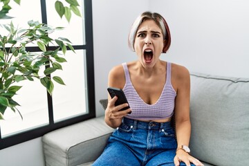 Young beautiful woman using smartphone typing message sitting on the sofa angry and mad screaming...