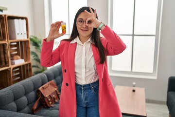 Young latin woman working as therapist holding jar of pills smiling happy doing ok sign with hand...
